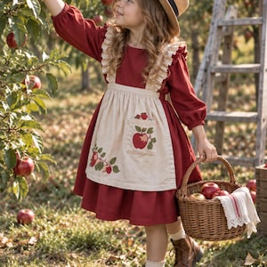 May include: A long-sleeved dark red dress with a beige apron featuring an apple print, paired with a straw hat, brown lace-up boots, and a woven wicker basket. The dress has ruffled shoulder straps and the apron includes a front pocket detail.
