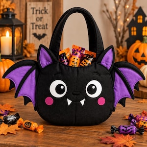 Könnte beinhalten: Eine schwarze Halloween-Süßigkeitentasche aus Stoff in Fledermausform mit großen weißen Augen, zwei weißen Fangzähnen, rosa Wangen und violetten Flügelinnenseiten. Die Tasche hat einen stabilen Griff und ist mit kleinen, verpackten Süßigkeiten gefüllt.