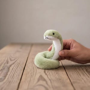 Könnte beinhalten: Ein Plüschschlangenspielzeug mit weichem, salbeigrünem Fell auf der Oberseite und einer weißen Unterseite. Das Stofftier hat einen geringelten Körper, einen abgerundeten Kopf und kleine schwarze Perlenaugen. Der Stoff ist flauschig und die Schlange aufgerichtet.