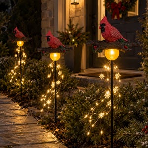 May include: Three outdoor garden stakes featuring red cardinal birds perched on textured branches. Each stake has a glowing amber orb light and cascading wire strings adorned with small, illuminated white snowflake shapes.