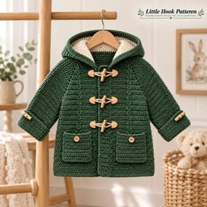 May include: A forest green hooded crochet coat featuring three light-coloured wooden toggle closures down the front, two front patch pockets with wooden buttons, and long sleeves with matching toggle details. The interior is lined with cream yarn.