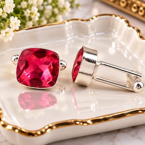 Può includere: Un paio di gemelli color argento con grandi pietre rosa acceso, tagliate a forma quadrata e sfaccettate. I gemelli presentano una finitura metallica lucida e un classico meccanismo di chiusura a perno, che riflette la luce sulle superfici.