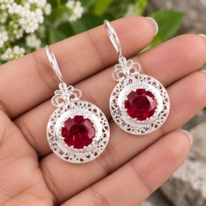 May include: A pair of silver drop earrings featuring a round, faceted deep red stone set in the center of a circular frame with intricate filigree metalwork and a decorative scroll-like top, suspended from a curved silver ear wire hook.