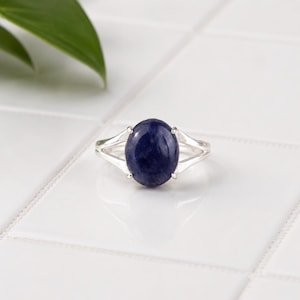 Peut inclure: Une bague en argent ornée d'une pierre précieuse bleu foncé de forme ovale, fixée par un sertissage à griffes. L'anneau de la bague se divise en deux branches distinctes au niveau de la pierre centrale, offrant un design épuré et symétrique.