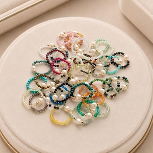 May include: A collection of circular rings crafted from small, multi-colored faceted beads and white pearls. Each ring features a single white pearl accented by tiny gold-toned metal beads, interspersed among various translucent gemstone-colored beads.