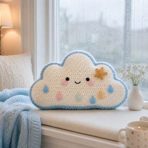 May include: A white crochet cloud pillow with a light blue border, featuring a smiling face with pink cheeks, black eyes, and a small yellow star. The cloud is decorated with several small, colorful raindrops in shades of blue, pink, and yellow.