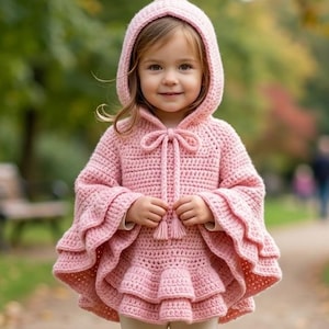 May include: A light pink crocheted hooded poncho featuring a tiered ruffled design on the sleeves and a drawstring closure at the neck with two tassels. The garment is paired with plain cream-colored leggings and brown leather ankle boots.