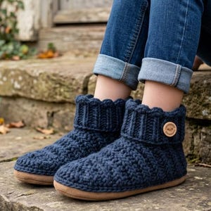 Könnte beinhalten: Ein Paar dunkelblaue Häkelhausschuhe mit einem dicken gerippten Bündchen, einem strukturierten Strickmuster und einem kleinen runden Holzknopf an der Seite. Die Hausschuhe haben eine flache, hellbraune Sohle und ein knöchelhohes Design.