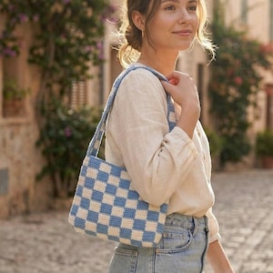 May include: A rectangular crochet shoulder bag featuring a blue and white checkered pattern. The bag has a matching blue and white crocheted strap and is worn over the shoulder. The construction shows a consistent, dense stitch pattern throughout.