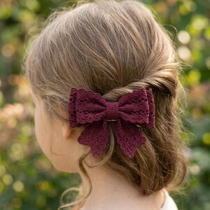 May include: A deep burgundy hair bow featuring a detailed lace-like texture with scalloped edges. The bow is constructed with a layered design, creating a structured appearance while secured to the hair, showcasing a rich, dark red color.