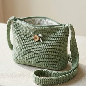 May include: A sage green crochet crossbody bag featuring a single wooden button closure at the centre, accented with a small matching ribbon bow. The bag has a zippered top opening and a light-coloured interior lining with a subtle leaf pattern.