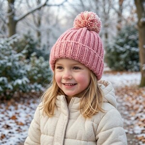 Op de afbeelding: Een oudroze gebreide muts met een dikke geribbelde textuur en een grote bijpassende pompon aan de bovenkant. De muts heeft een brede omgeslagen boord aan de onderkant en is gemaakt van grof garen, wat zorgt voor een warme uitstraling.