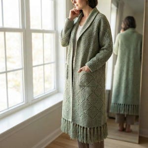 May include: A sage green long-line cardigan featuring a textured diamond pattern, a shawl collar, two front patch pockets, and a long fringe trim along the bottom hem. The knit garment has long sleeves and an open front design.
