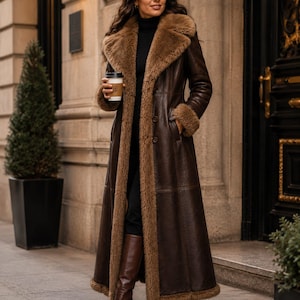 May include: A long, dark brown leather coat featuring a thick, tan faux-fur lining along the wide lapels, cuffs, and hem. The coat is fastened with two visible buttons down the front and is worn over a black turtleneck sweater and brown boots.