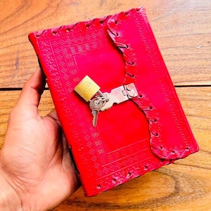 May include: A bright red leather journal featuring an embossed geometric border pattern and a stitched edge detail. The journal is secured with a silver-toned metal latch and a small brass padlock, which includes a single key attached to the mechanism.