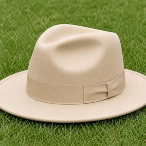 Könnte beinhalten: Ein beigefarbener Fedora-Hut mit einer strukturierten Krone und einer breiten, flachen Krempe. Der Hut verfügt über ein passendes beiges Ripsband, das die Basis der Krone umschließt und ein kleines, gefaltetes Schleifendetail an der Seite aufweist.