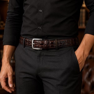 May include: A dark brown belt with a textured crocodile skin pattern and a silver-toned metal buckle. The belt is worn around the waist over black trousers, paired with a black button-down shirt. The design features a classic rectangular buckle.