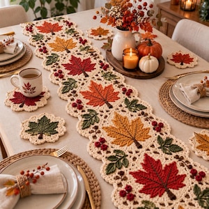 May include: A cream-coloured crochet table runner featuring a repeating pattern of red, orange, and yellow maple leaves interspersed with small clusters of red berries and green leaves, accompanied by matching circular crochet coasters.