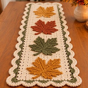 Pode incluir: Um caminho de mesa retangular de crochê em fio creme com uma borda festonada em verde escuro. O caminho apresenta quatro grandes aplicações de folhas de bordo texturizadas em cores alternadas: amarelo mostarda, laranja queimado e verde oliva.