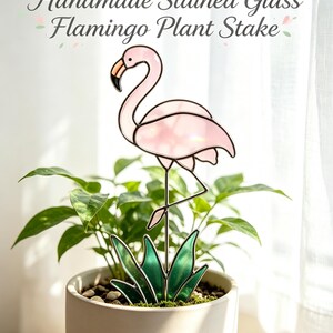 Puede incluir: Un tutor para plantas de vidrio tintado hecho a mano con un flamenco rosa claro con pico negro y naranja sobre follaje de vidrio verde. El texto dice: Handmade Stained Glass Flamingo Plant Stake, For Potted Plants & Garden Soil, Plant Lover Gift, Tropical Garden Decor.