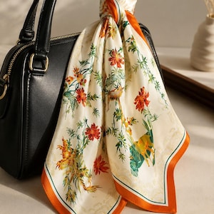 May include: A black leather handbag with a square silhouette and gold-tone hardware, featuring a cream-colored square scarf tied to the handle. The scarf displays a detailed print of orange flowers, green foliage, and deer in a natural setting.