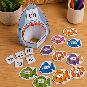 May include: A paper shark cutout with an open mouth featuring the letters ch, surrounded by various colorful fish cutouts labeled with words like fish, moth, chin, dish, chip, chop, path, quiz, quack, and quit, plus letter tiles th, ee, sh, ng, and ai.