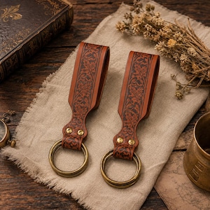 May include: Two tan leather belt loops featuring an intricate embossed maple leaf pattern, secured with two brass rivets and attached to a circular brass ring. The leather is dyed a medium brown with darker accents highlighting the leaf design.
