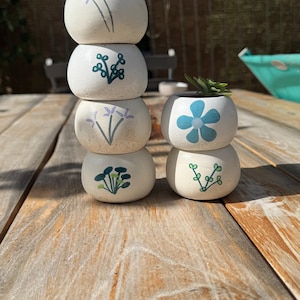 Peut inclure: Un ensemble de six petits pots de fleurs en céramique blanc cassé, de forme sphérique, disposés en deux piles. Les pots présentent des motifs botaniques peints à la main en bleu, vert et violet, incluant des fleurs et des tiges feuillues.