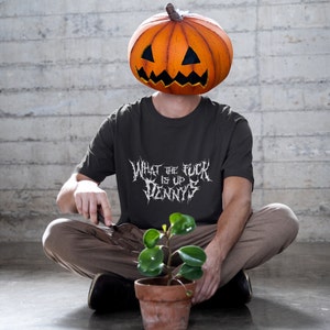 May include: A person wearing a dark grey t-shirt with the text "What the f*ck is up Dennis" in white metal font. The person is wearing a carved pumpkin on their head. The person is sitting on the floor with a potted plant in front of them.