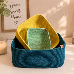 May include: A set of three nested square storage baskets made of coiled rope. The largest basket is dark teal, the medium basket is bright yellow, and the smallest basket is light green. Each basket features a small brown leather-like side tab.