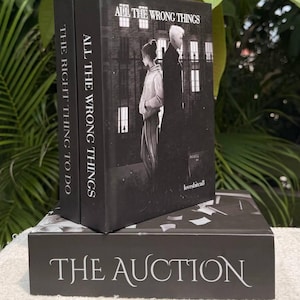 May include: A stack of three black books with white text. The top book is titled All The Wrong Things, featuring an illustration of two figures. The spine of the middle book reads All The Wrong Things, the bottom book is titled The Auction.