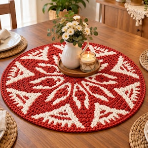 May include: A round, crocheted table centrepiece featuring a bold red and white geometric snowflake pattern. The circular mat is centred on a wooden table, topped with a white ceramic pitcher filled with white daisies and a lit candle in a glass jar.