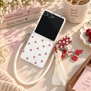 May include: A white foldable smartphone case featuring a repeating red cherry pattern, equipped with a long white wrist strap and a decorative charm cluster including red beads, a white star, and a red heart attached to the side.
