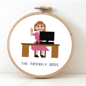 Op de afbeelding: Een borduurring met een wit achtergrond waarop een gepixelde afbeelding van een vrouw te zien is die aan een bureau met een computer zit. De vrouw draagt een roze jurk en heeft bruin haar. De tekst "THE FRIENDLY BOSS" staat onder de afbeelding.