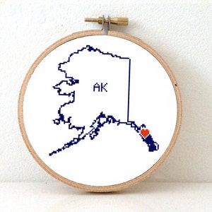 May include: Embroidered hoop art featuring a blue outline of the state of Alaska with a red heart over the city of Anchorage. The state abbreviation "AK" is written in the center of the state.