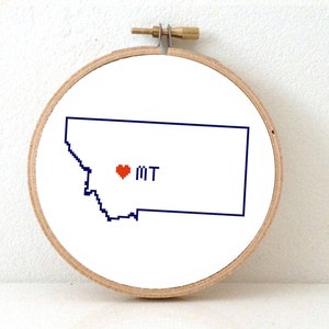 May include: A white embroidery hoop with a blue outline of the state of Montana and a red heart with the letters "MT" inside.