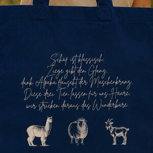 May include: A dark blue tote bag featuring a light-colored graphic print. The design includes a five-line German poem in cursive script above illustrations of an alpaca, a sheep, and a goat, all rendered in a simple, clean line-art style.