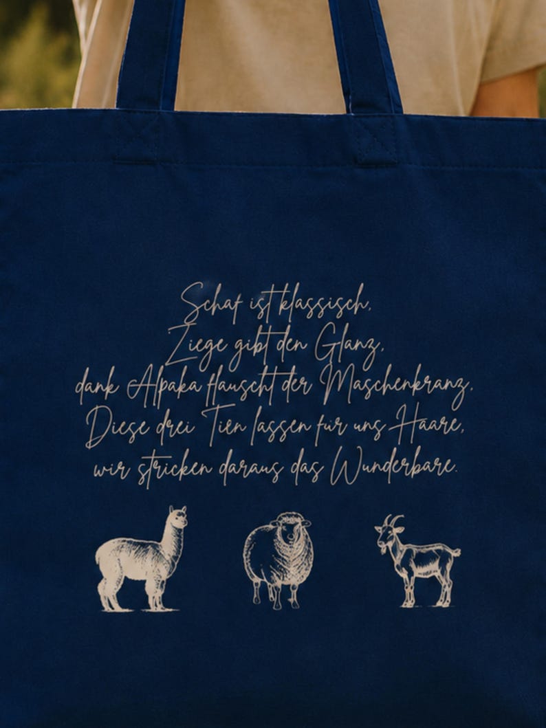 May include: A dark blue tote bag featuring a light-colored graphic print. The design includes a five-line German poem in cursive script above illustrations of an alpaca, a sheep, and a goat, all rendered in a simple, clean line-art style.