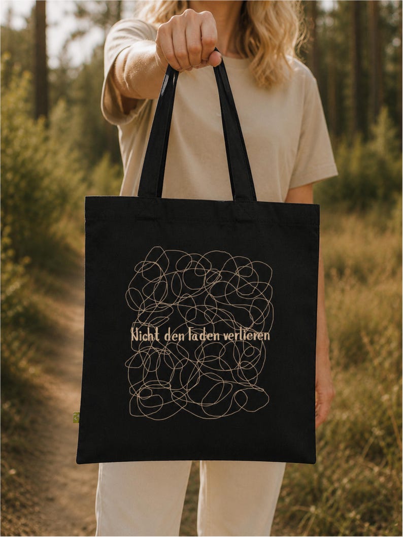 May include: A black tote bag featuring a beige line art graphic of a tangled, chaotic scribble pattern. Centered within the design is the text "Nicht den Faden verlieren" printed in a beige, serif-style font. The bag has two black shoulder straps.