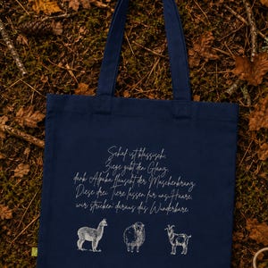 May include: A navy blue tote bag featuring a white line-art illustration of an alpaca, a sheep, and a goat. Above the animals, the bag displays German text that reads: Schaf ist klassisch, Ziege gibt den Glanz, dank Alpaka hauscht der Maschenkranz.