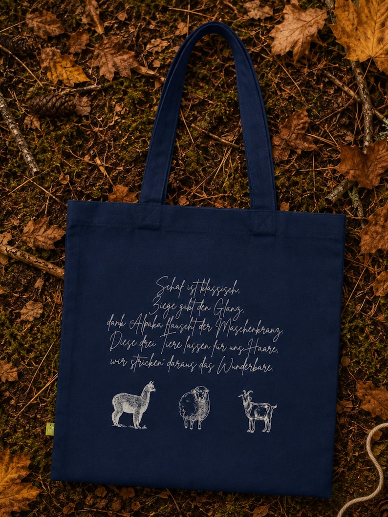 May include: A navy blue tote bag featuring a white line-art illustration of an alpaca, a sheep, and a goat. Above the animals, the bag displays German text that reads: Schaf ist klassisch, Ziege gibt den Glanz, dank Alpaka hauscht der Maschenkranz.