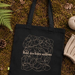 May include: A black tote bag featuring a beige line art design of tangled yarn and the German text "Nicht den Faden verlieren" printed in the center. The bag has two long shoulder straps and a small green tag on the lower left side.
