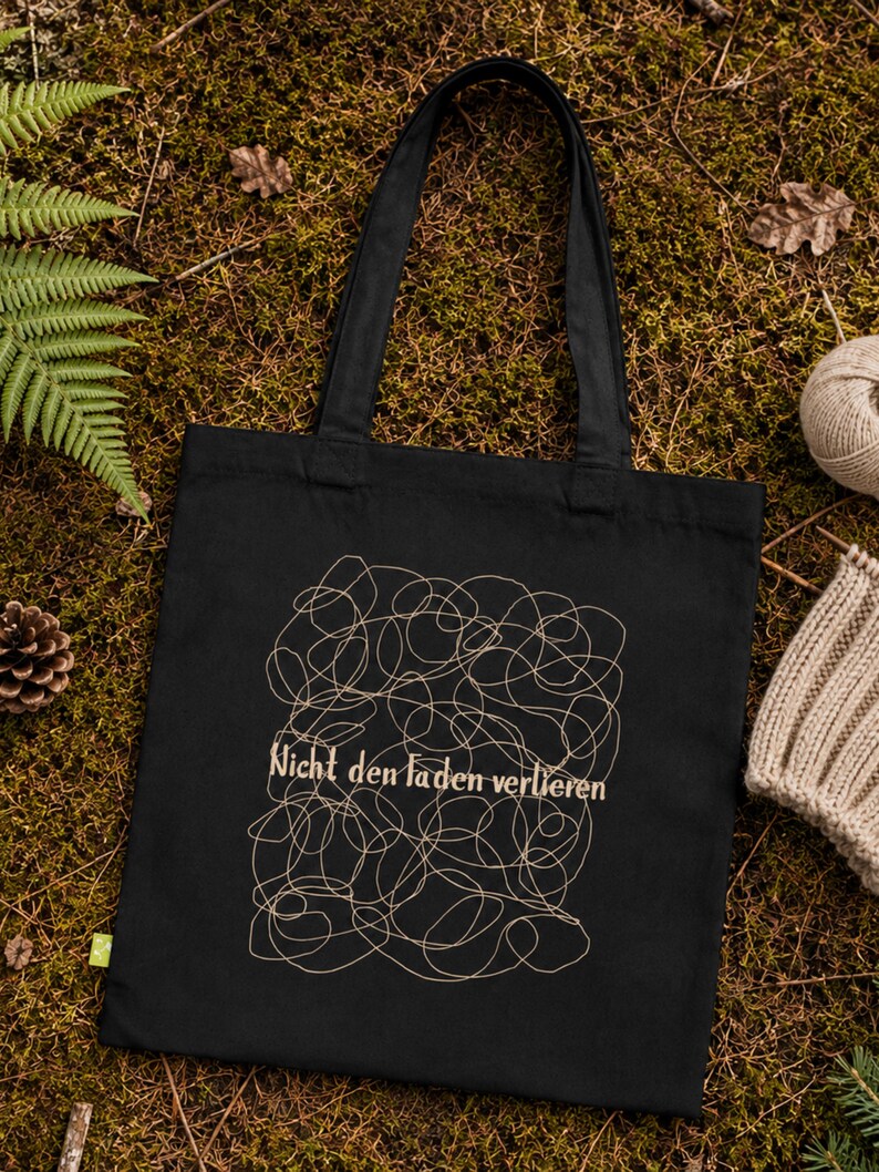 May include: A black tote bag featuring a beige line art design of tangled yarn and the German text "Nicht den Faden verlieren" printed in the center. The bag has two long shoulder straps and a small green tag on the lower left side.