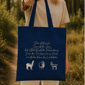 May include: A navy blue tote bag featuring a white graphic illustration of an alpaca, a sheep, and a goat. Above the animals, the bag displays German text that reads: Schaf ist klassisch. Ziege gibt den Glanz, dank Alpaka haucht der Maschenkrang. Diese drei Tier lassen f&uuml;r uns Haare, wir stricken daraus das Wunderbare.