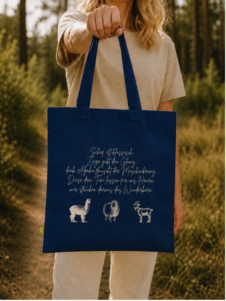 May include: A navy blue tote bag featuring a white graphic illustration of an alpaca, a sheep, and a goat. Above the animals, the bag displays German text that reads: Schaf ist klassisch. Ziege gibt den Glanz, dank Alpaka haucht der Maschenkrang. Diese drei Tier lassen f&uuml;r uns Haare, wir stricken daraus das Wunderbare.
