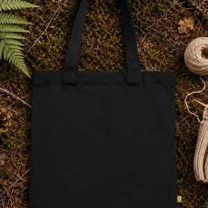 May include: A solid black rectangular tote bag with two matching fabric shoulder straps. The bag features a small, light green rectangular tag attached to the lower right side seam, providing a subtle contrast against the plain black fabric.