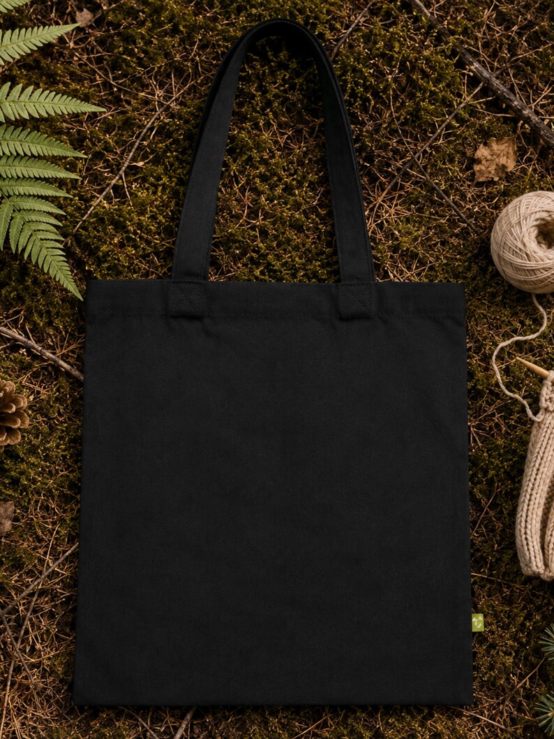 May include: A solid black rectangular tote bag with two matching fabric shoulder straps. The bag features a small, light green rectangular tag attached to the lower right side seam, providing a subtle contrast against the plain black fabric.