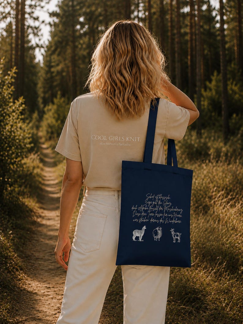 May include: A beige t-shirt with the text COOL GIRLS KNIT and a dark blue tote bag featuring white illustrations of an alpaca, a sheep, and a goat, accompanied by German text printed in a script font on the front of the bag.