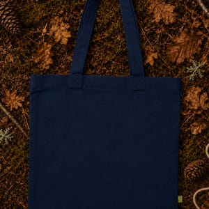 May include: A solid navy blue canvas tote bag with two matching fabric shoulder straps. The bag features a rectangular body with a flat base and a small green tag attached to the lower right side seam, presenting a clean and simple design.