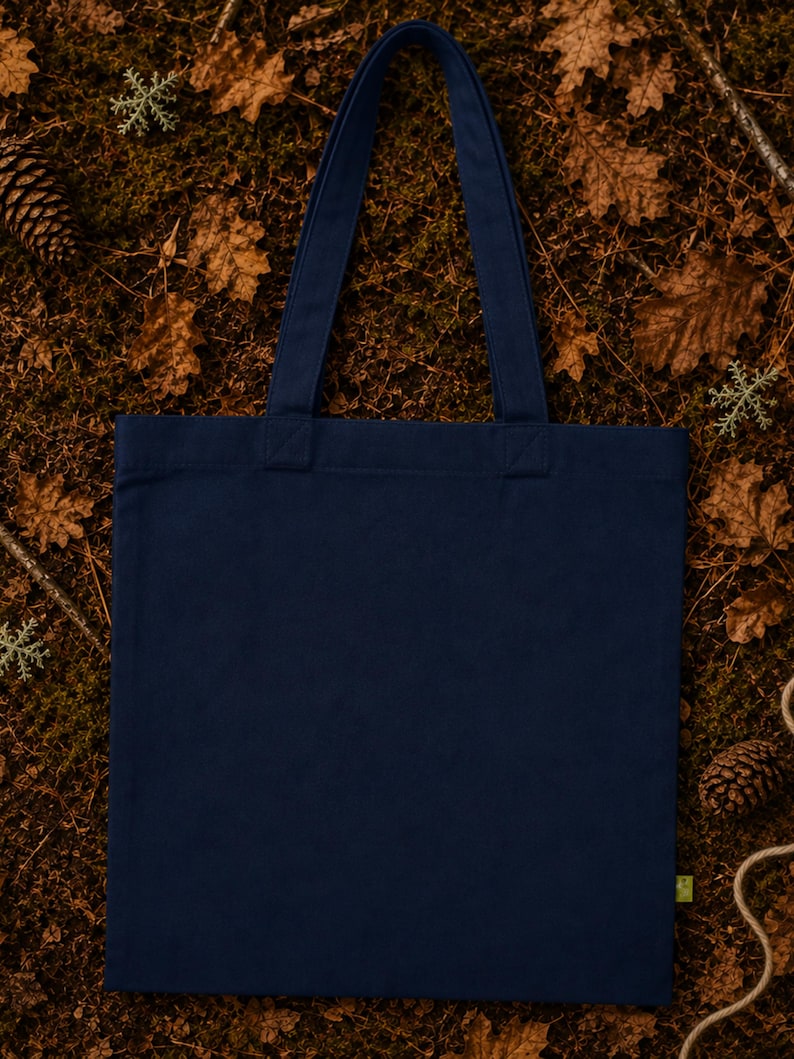 May include: A solid navy blue canvas tote bag with two matching fabric shoulder straps. The bag features a rectangular body with a flat base and a small green tag attached to the lower right side seam, presenting a clean and simple design.